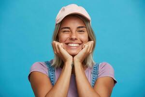 fechar-se do sorridente bonita Loiras jovem mulher desgasta Rosa boné e tolet t camisa parece feliz e piscando isolado sobre azul fundo foto