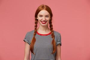 menina com dois vermelho cabelos tranças, encantador cheio de dentes sorriso, usava vermelho batom vestido dentro despojado camiseta, isolado em uma Rosa estúdio fundo foto