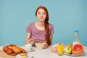 fofa ruivo menina com uma trança senta às uma mesa, tem café da manhã, com excitação come flocos de milho com leite, parece às Câmera com tentação, doces em a mesa e fresco fruta, em uma azul fundo foto