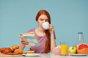 cutelo ruivo menina com trançado cabelo, sentado às uma mesa, bebidas a partir de branco copo delicioso chá, tem café da manhã lendo livro. em mesa cozimento produtos e fresco Comida escala azul parede foto