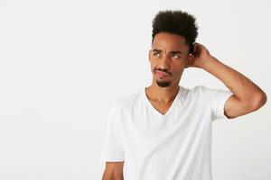 retrato do pensativo sério africano americano jovem homem com encaracolado cabelo desgasta t camisa parece pensativo e coçar dele cabeça isolado sobre branco fundo foto