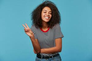 africano americano bonita sorridente menina com afro Penteado olhando às Câmera e fazendo Paz placa com mão ou v placa com dedos. isolado sobre azul fundo, cópia de espaço foto