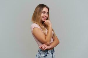 retrato do atraente, adulto menina com loiro grandes cabelo. vestindo Rosa camiseta e jeans. tocante dela queixo e sorriso. assistindo às a Câmera sobre dela ombro, isolado sobre cinzento fundo foto