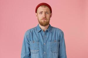 retrato do de olhos azuis jovem cara dentro vermelho chapéu com vermelho barba parece triste, chateado, frustrado, descontente com algo, lábios beicinho, expressa insulto, desgasta elegante jeans camisa, isolada em Rosa fundo foto