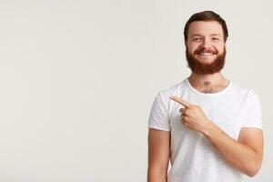 fechar-se do alegre confiante jovem homem hipster com barba desgasta t camisa sente feliz e pontos para a lado com dedo isolado sobre branco fundo foto