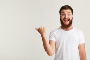 fechar-se do alegre animado jovem homem hipster com barba desgasta t camisa sente feliz e pontos para a lado às copyspace com dedo isolado sobre branco fundo foto