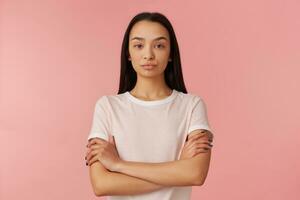 retrato do sério, descontente menina com Preto grandes cabelo. vestindo branco camiseta. pessoas e emoção conceito. mantém braços cruzado, esperando. assistindo às a Câmera isolado sobre pastel Rosa fundo foto