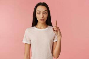 chocado olhando mulher, lindo menina com grandes Preto cabelo. vestindo branco camiseta. levanta dedo acima. lembrar, aviso prévio algo. assistindo às a Câmera isolado sobre pastel Rosa fundo foto