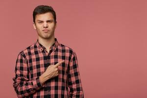 retrato do franzindo a testa alegre cara desgasta dentro xadrez camisa, parece às a Câmera com com nojo expressão, quer para virar seu atenção para cópia de espaço às a certo lado, carrinhos sobre Rosa fundo. foto