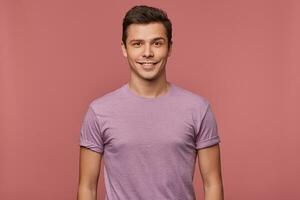 foto do jovem atraente homem dentro blanc camiseta, alegre parece às a Câmera, carrinhos sobre Rosa fundo e sorri.