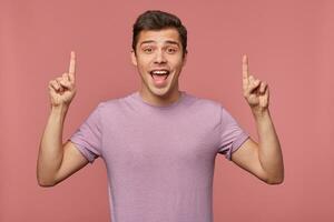 retrato do jovem atraente feliz espantado homem desgasta dentro em branco camiseta, parece acima e quer para virar seu atenção para cópia de espaço sobre dele cabeça , isolado sobre Rosa fundo. foto