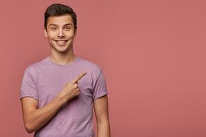 jovem bonito alegre homem desgasta dentro em branco camiseta, parece às a Câmera com feliz expressão, quer para virar seu atenção para cópia de espaço às a certo lado, carrinhos sobre Rosa fundo. foto