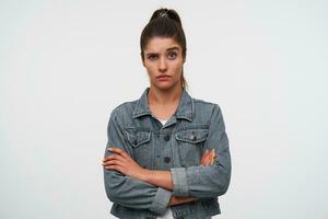 retrato do jovem morena senhora com elevado sobrancelha desgasta dentro branco camiseta e jeans jaquetas, parece às a Câmera com infeliz expressão, carrinhos sobre branco fundo com cruzado braços. foto