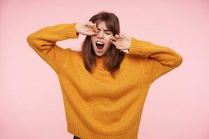 cansado jovem atraente morena mulher dentro acolhedor de lã guardando dela olhos fechadas enquanto bocejando com Largo boca aberto, levantando mãos para dela face enquanto posando sobre Rosa fundo foto