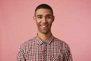 foto do jovem alegre atraente Sombrio esfolado cara dentro xadrez camisa, parece às a Câmera com feliz expressão e amplamente sorridente, carrinhos sobre Rosa fundo.