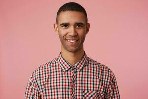 retrato do jovem feliz atraente Sombrio esfolado cara dentro xadrez camisa, parece às a Câmera com positivo expressão, amplamente sorridente e carrinhos sobre Rosa fundo, positivo emoção conceito. foto