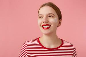 fechar acima do atraente jovem sorridente ruivo mulher com vermelho lábios e com remendos debaixo a olhos, desgasta dentro uma vermelho listrado camiseta, parece longe com uma feliz expressão, carrinhos sobre Rosa fundo. foto