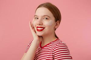 jovem sorridente ruivo menina com vermelho lábios e com remendos debaixo a olhos, desgasta dentro uma vermelho listrado camiseta, toques a bochecha, carrinhos sobre Rosa fundo e desfrutando livre Tempo para pele Cuidado. foto