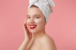 fechar acima do jovem sorridente mulher com uma toalha em dela cabeça depois de banho, com remendos e vermelho lábios, toques face e parece feliz, carrinhos sobre Rosa fundo. foto