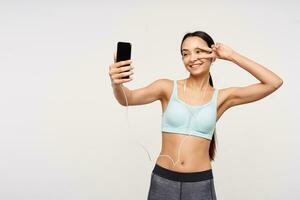 alegre jovem bonita Castanho cabelos senhora com rabo de cavalo Penteado levantando mão com vitória gesto para dela face enquanto fazer selfie em Móvel telefone, em pé sobre branco fundo foto