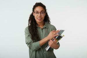 retrato do alegre jovem encaracolado morena Sombrio esfolado senhora com casual Penteado guardando livros didáticos dentro elevado mãos e sorridente amplamente às Câmera, isolado sobre branco fundo foto