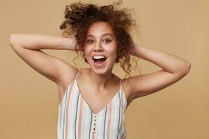 alegre jovem atraente mulher guardando dela encaracolado Castanho cabelo com elevado mãos e olhando alegremente às Câmera com Largo sorriso, posando sobre bege fundo dentro verão topo foto