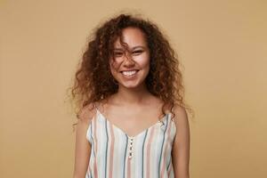 retrato do alegre jovem bonita encaracolado morena mulher olhando às Câmera com encantador sorrir e guardando dela mãos baixa enquanto em pé sobre bege fundo foto