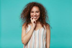 retrato do jovem olhos castanhos encaracolado morena mulher com natural Maquiagem guardando índice dedo em dela lábio inferior e olhando às Câmera com Largo sincero sorriso, isolado sobre azul fundo foto