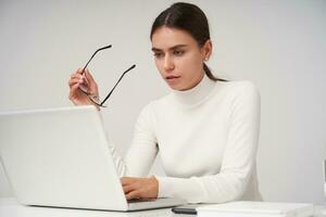 coletado jovem lindo morena mulher com rabo de cavalo Penteado segurando óculos dentro elevado mão enquanto olhando às dela computador portátil com concentrado face, sentado sobre branco fundo foto