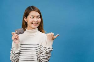 jovem senhora asiática mostra cartão de crédito com expressão positiva, sorri amplamente, vestida com roupas casuais, sentindo felicidade e carrinho isolado sobre fundo azul. conceito de expressão facial. foto