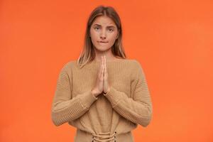 confuso jovem bonita cabeça branca fêmea com natural Maquiagem levantando mãos com Rezar gesto e morder preocupantemente lábio inferior enquanto em pé sobre laranja fundo foto