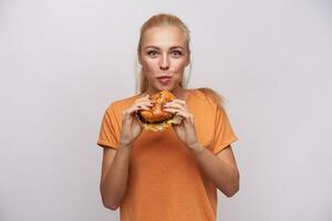 interior foto do atraente jovem com fome Loiras senhora com rabo de cavalo Penteado mastigar delicioso Hamburger e olhando excitadamente às Câmera, vestido dentro laranja camiseta sobre branco fundo