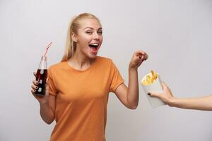 alegre jovem atraente Loiras fêmea com rabo de cavalo Penteado guardando garrafa com Palha dentro elevado mão e alcançando para francês fritas com Largo feliz sorriso, isolado sobre branco fundo foto