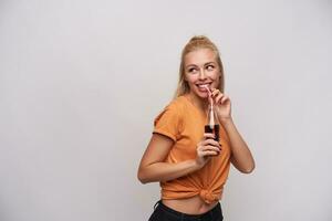 estúdio foto do alegre jovem atraente mulher dentro laranja camiseta bebendo refrigerante com Palha enquanto olhando a parte, de lado com encantador sorriso, posando contra branco fundo