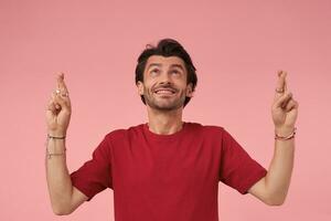 retrato do jovem bonito homem com na moda corte de cabelo em pé sobre Rosa fundo dentro casual roupas, levantando mãos com cruzado dedos para Boa sorte e olhando positivamente para cima foto