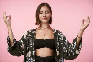 agradável olhando jovem positivo morena mulher levantando mãos dentro namaste gesto e sorridente levemente enquanto em pé sobre Rosa fundo dentro floresceu camisa foto