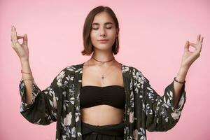 Boa olhando jovem calma curto cabelos morena senhora guardando dela olhos fechadas enquanto meditando com elevado mãos, isolado sobre Rosa fundo dentro na moda roupas foto