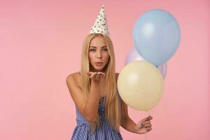 retrato do atraente jovem grandes cabelos mulher a comemorar aniversário com multicolorido ar balões, olhando às Câmera positivamente e sopro ar beijo com franzido lábios foto
