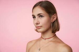 retrato do jovem lindo olhos castanhos mulher com prumo corte de cabelo guardando dela lábios guardada enquanto olhando suavemente aparte, em pé sobre Rosa fundo com mãos baixa foto