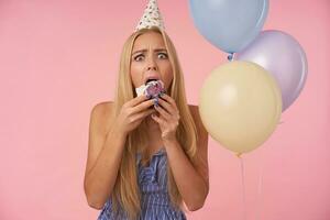 interior foto do jovem bonita fêmea com grandes Loiras cabelo vestindo azul verão vestir e aniversário boné, ser surpreso enquanto comendo bolo e olhando às Câmera com Largo olhos aberto