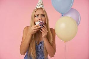 espantado jovem Loiras mulher dentro festivo roupas e cone chapéu franzindo a testa dela sobrancelhas e arredondamento olhos enquanto comendo delicioso torta dentro colori hélio balões, isolado sobre Rosa fundo foto