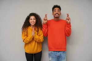 estúdio tiro do na esperança atraente jovem Sombrio esfolado encaracolado par vestindo de lã colorida blusas e jeans enquanto em pé sobre cinzento fundo com elevado mãos e sorridente amplamente foto