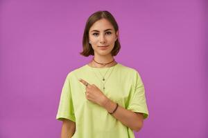 fêmea estudante, jovem senhora com morena curto cabelo, apontando dela dedo para a esquerda às a cópia de espaço sobre roxa fundo. mostrando uma placa. vestindo verde camiseta, pulseiras e argolas foto