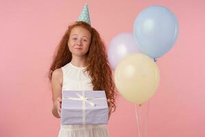 foto do fofa pequeno menina com grandes foxy cabelo ser divertido de convidados, sorrisos alegremente, isolado sobre Rosa estúdio fundo com ar balões, segurando aniversário apresenta e olhando para Câmera alegremente