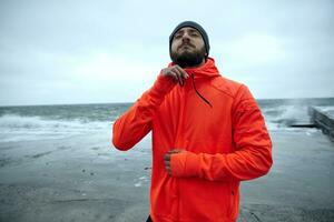 ao ar livre foto do jovem morena masculino com exuberante barba em pé sobre mar Visão em tormentoso cinzento dia e zipando acima dele caloroso laranja desportivo casaco. ginástica e esporte conceito.