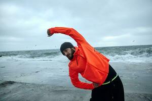 imagem do jovem Sombrio cabelos desportivo homem com barba fazendo fisica exercícios sobre beira-mar fundo em cinzento frio manhã, alongamento músculos antes dar certo. ginástica masculino modelo foto