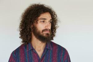 retrato do bonita jovem morena encaracolado homem com barba guardando lábios guardada enquanto olhando a parte, de lado com calma face, vestindo listrado multicolorido camisa enquanto posando contra branco fundo foto