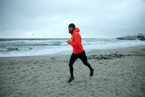 tiro do jovem barbudo Atlético masculino vestido dentro caloroso desportivo roupas corrida em litoral do mar dentro cedo cinzento manhã, olhando adiante com concentrado face e guardando mãos às peito nível foto