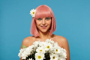 romântico foto do jovem adorável Rosa cabelos mulher com colori Maquiagem segurando enorme ramalhete do flores e olhando positivamente às Câmera com encantador sorriso, isolado sobre azul fundo