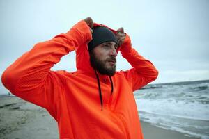 retrato do jovem atraente morena barbudo homem dentro caloroso desportivo roupas colocando em de capuz enquanto em pé sobre litoral do mar depois de manhã corrida sessão foto